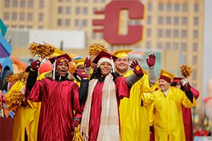 A group of people in maroon and gold graduation gowns and caps waving at parade-goers.