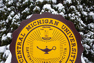 The Central Michigan University seal in front of a snow-covered bush.