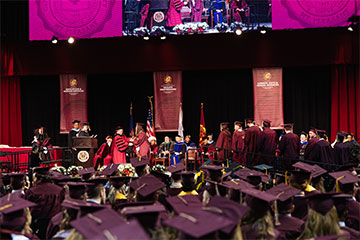 Graduates wearing maroon caps and gowns watch a commencement ceremony onstage.