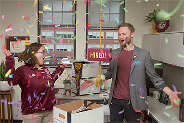 A young male and female stand in an office as confetti falls from the ceiling. A yellow rope with a sign reading "hired" hangs between them.