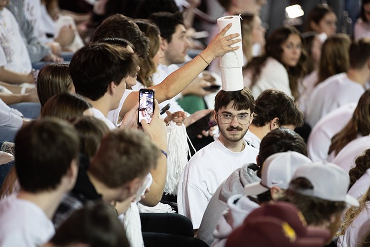 In a large crowd at a CMU men's basketball games, a student stares off into the distance as another student balances three rolls of toilet paper on his head.