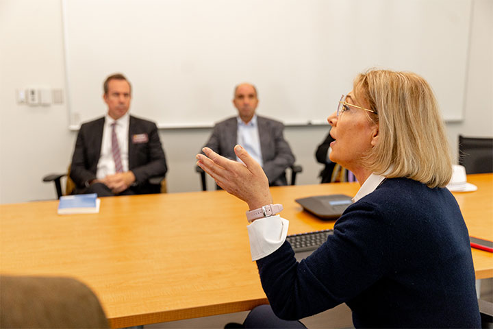 A blonde woman in a navy sweater sits across a wooden desk from two men in suits