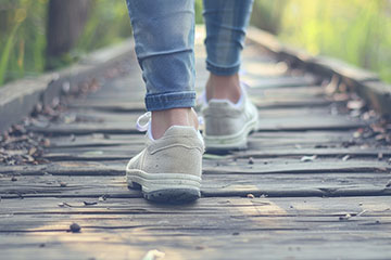 A pair of feet in white sneakers and attached to legs in denim pants walks down a wood boardwalk through grass.