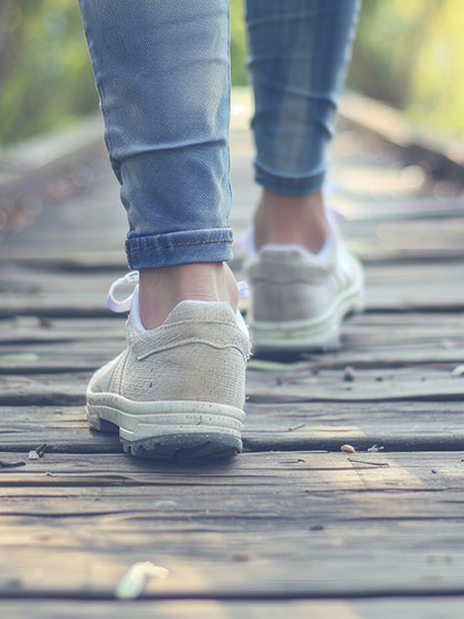 A pair of feet in white sneakers and attached to legs in denim pants walks down a wood boardwalk through grass.