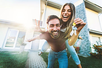 A bearded smiling man holds keys to a house in the background while a smiling woman with long hair balances on his back.