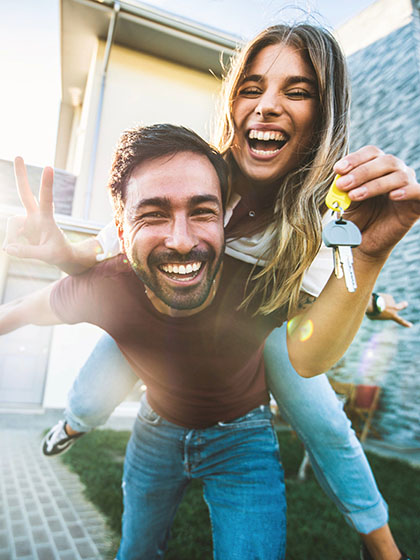 A bearded smiling man holds keys to a house in the background while a smiling woman with long hair balances on his back.