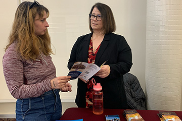 A woman with long hair holds a pamphlet while a second woman with medium-length hair explains its contents.