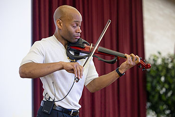 A man in a white shirt holds a violin with his left hand and chin while pulling a string across it with his right hand.