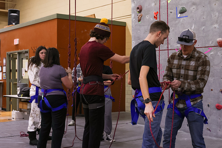 A group of students stand near a rock climbing wall as a CMU faculty member clips them into their lines for safety.