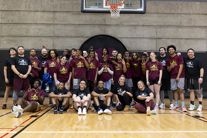 A group of approximately 20 students and staff pose for a photo on a basketball court inside the SAC.