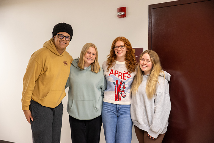 Four students stand together for a group photo while standing in front of a white wall and a door.