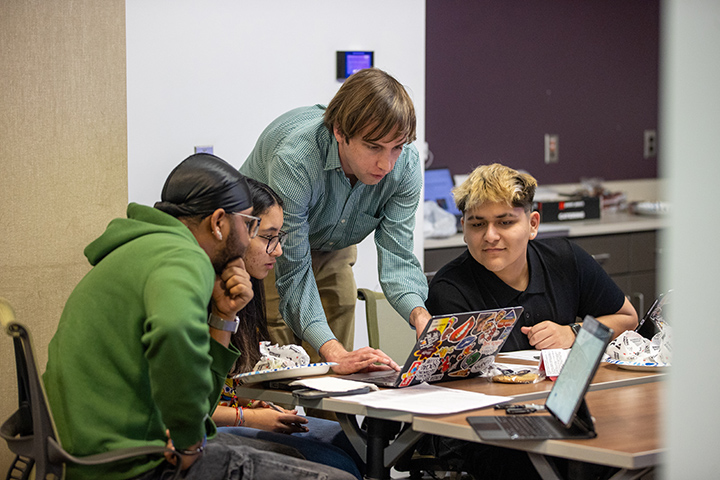 Four students huddle around a laptop at a table.