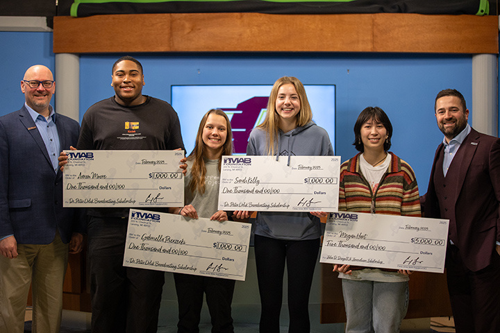 Four students hold large novelty checks as two men flank the group on either side.