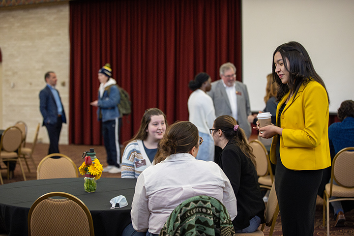 A female student in a yellow sweater stands next to a table talking to a group of students who are seated.