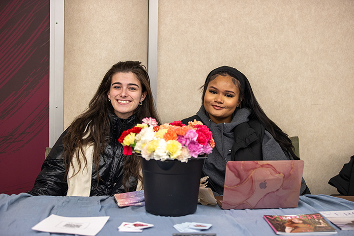 Two college students sit at a table. Between them is a bucket of flowers and a laptop computer.