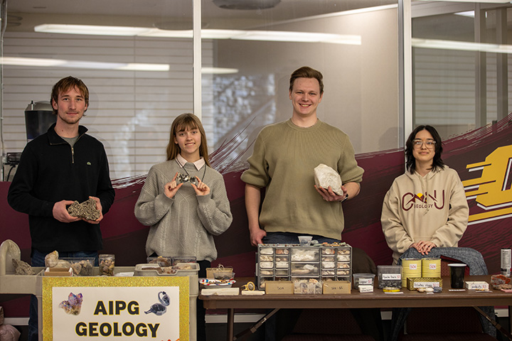 Four students stand behind a table smiling for the camera. On the table, a sign reads AIPG Geology.