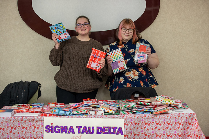 Two students stand behind a table holding up gift-wrapped books. A sign on the table reads Sigma Tau Delta.