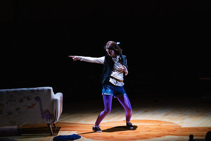 A theatre student stands in the spotlight on stage making a dance pose and pointing to the left.