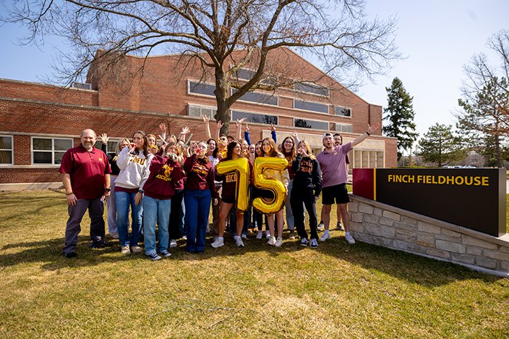 A group of 25 students and faculty stand next to the Finch Fieldhouse sign holding gold number balloons with the number 75.
