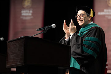 A man wearing black and green commencement regalia claps his hands