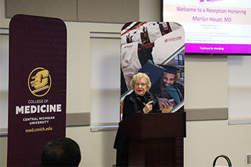 A woman in a black blazer stands behind a podium with a microphone near a banner reading College of Medicine