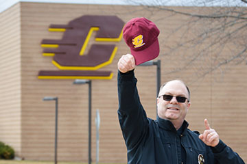 A police officer in uniform holds a maroon baseball hat in the air in front of the CMU logo