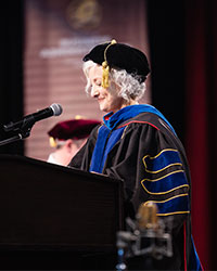 A woman in academic regalia stands at a podium at a graduation ceremony.