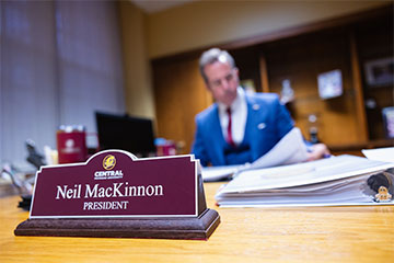 A maroon nameplate with the words Neil MacKinnon president sits on a desk in front of a man in a blue suit