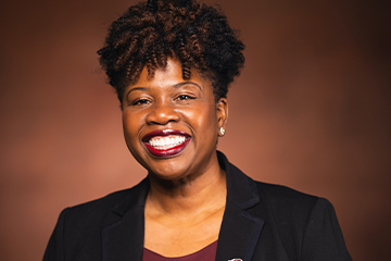 A woman with brown curly hair wearing a black blazer smiles at the camera.