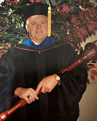 A man wearing a black academic cap and gown holds a commencement mace