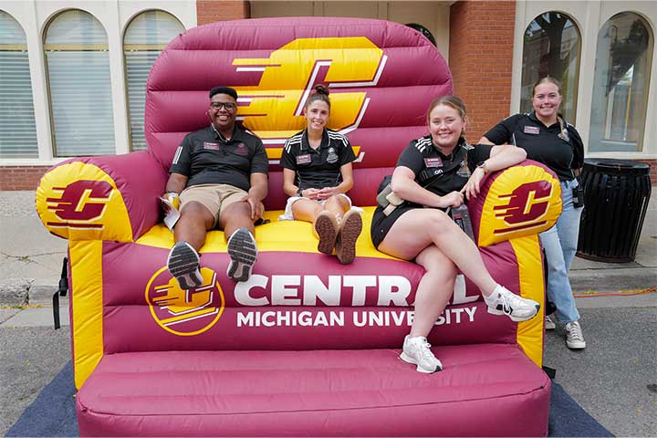 Four young people wearing black polo shirts sit on a giant maroon and gold inflatable chair.