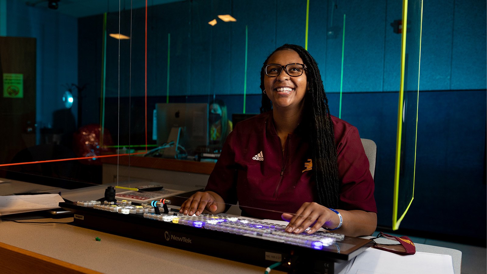 A female student in a maroon polo shirt sits in a dark broadcast studio lab, the keyboard lights up different colors.