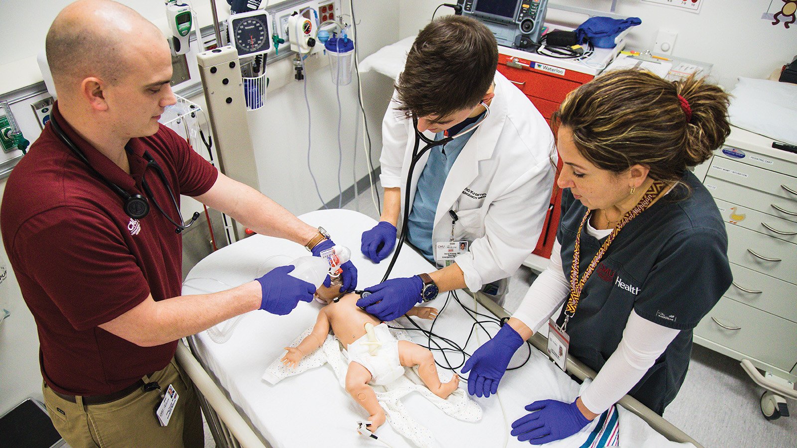 Medical students wearing blue gloves in a lab setting work with a medical simulator.