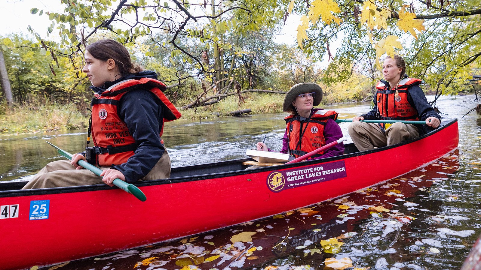 Three females wearing red orange life jackets with research equipment row down a river inside a red kayak