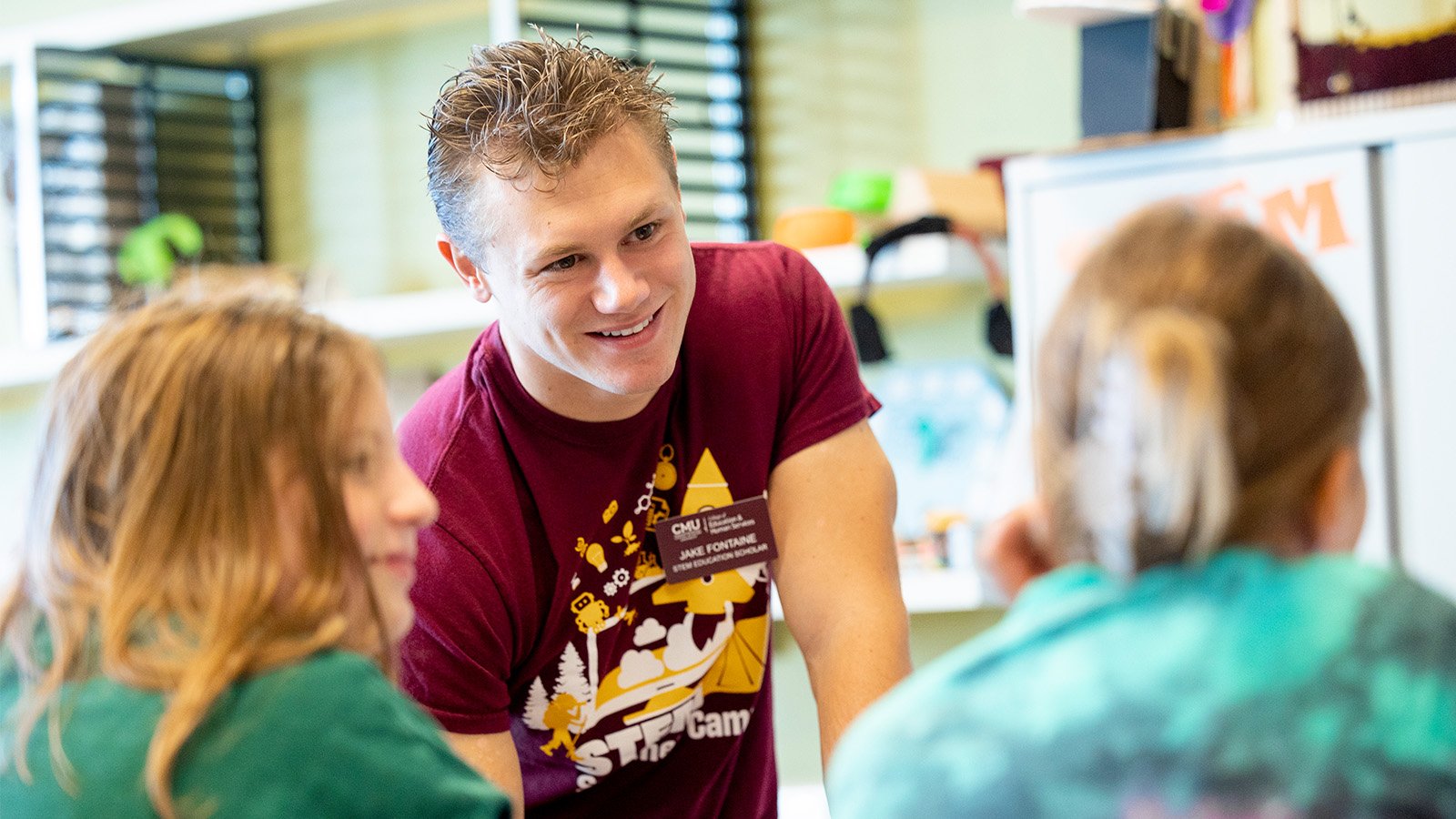 A male student teacher in a maroon shirt works with two female students in a STEM classroom.