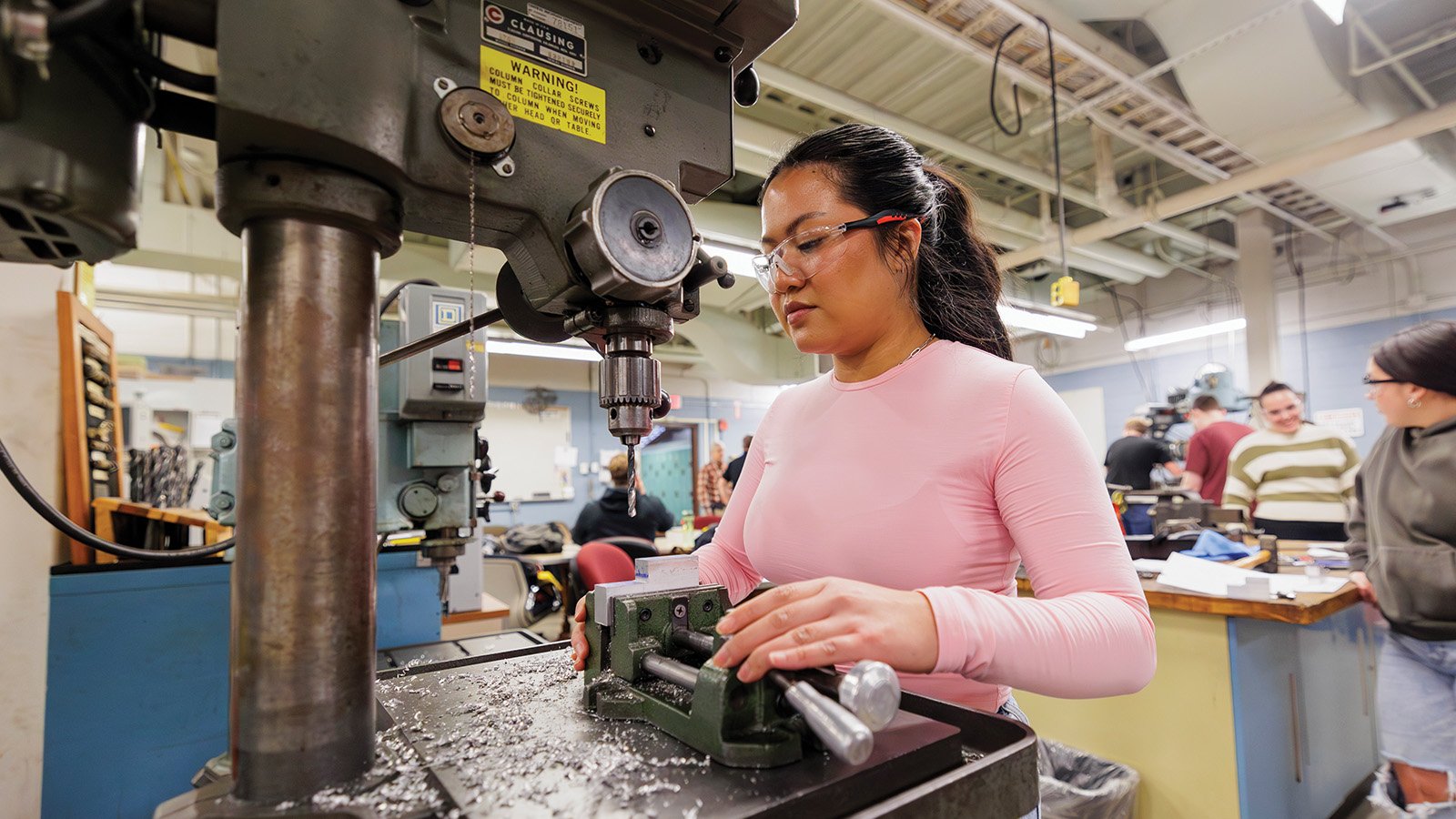 Female student wearing protective glasses stands at a machine inside an engineering lab.