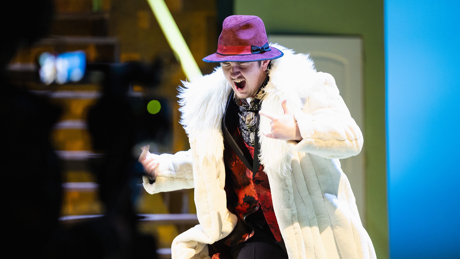 A male student performing on stage wearing a faux fur lined coat and a bright purple hat.