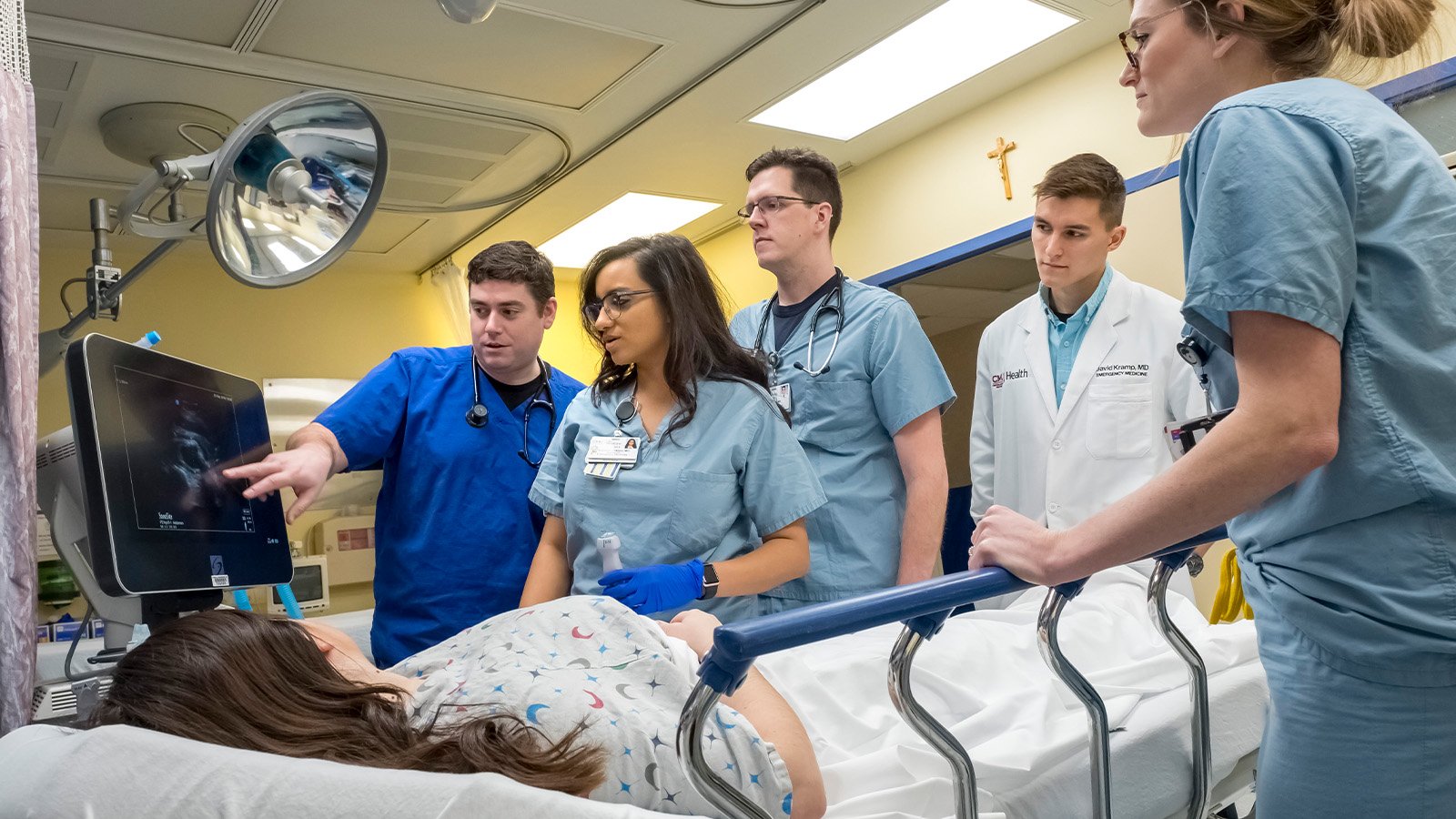 Medical students stand around a patient in a hospital bed while they review data on a computer monitor.