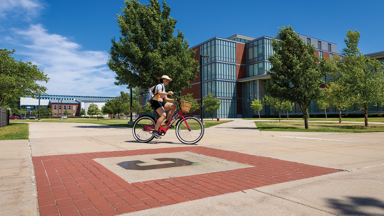 A female rides a red bike across campus in the summer, passing in front of a large bioscience building.