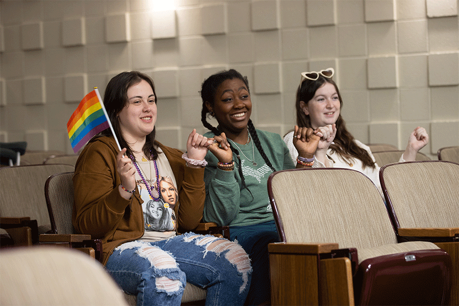 Three students are sitting in an auditorium, holding pinkies with each other as they attend an LGBTQ  event, and the student farthest to the left is holding a small pride flag.