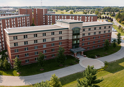 Aerial view of Campbell Hall on the campus of Central Michigan University, captured from a drone. The residence hall is surrounded by lush green grass and trees, with clear blue skies above.