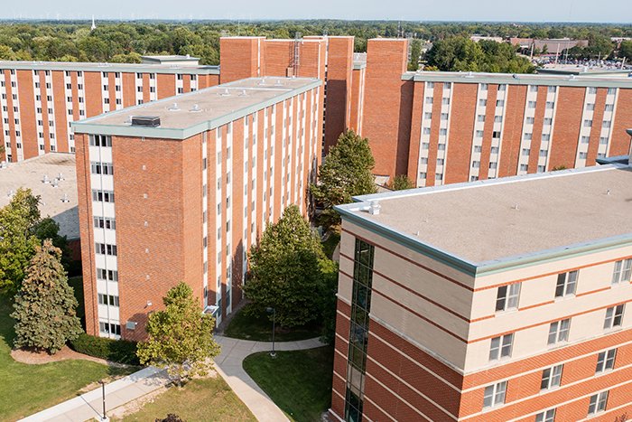 Aerial view of Carey Hall on the campus of Central Michigan University, captured from a drone. The residence hall is surrounded by lush green grass and trees, with clear blue skies above.