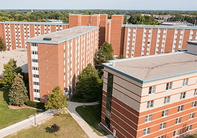 Aerial view of Carey Hall on the campus of Central Michigan University, captured from a drone. The residence hall is surrounded by lush green grass and trees, with clear blue skies above.