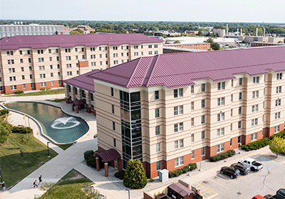 Aerial view of Celani Hall on the campus of Central Michigan University, captured from a drone. The residence hall is surrounded by lush green grass and trees, with clear blue skies above and a fountain outside.