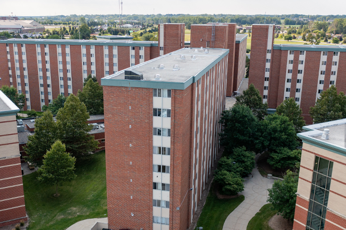 Aerial view of Cobb Hall on the campus of Central Michigan University, captured from a drone. The residence hall is surrounded by lush green grass and trees, with clear blue skies above.