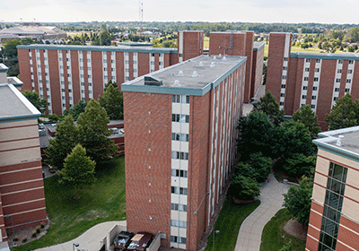 Aerial view of Cobb Hall on the campus of Central Michigan University, captured from a drone. The residence hall is surrounded by lush green grass and trees, with clear blue skies above.