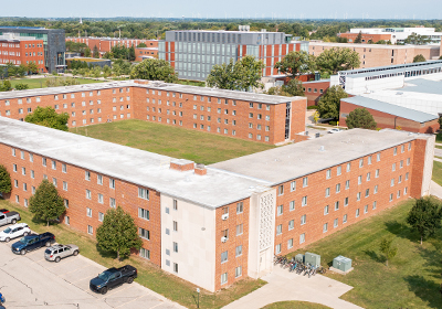 Aerial view of Emmons Hall on the campus of Central Michigan University, captured from a drone. The residence hall is surrounded by lush green grass and trees, with clear blue skies above.