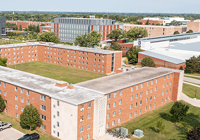 Aerial view of Emmons Hall on the campus of Central Michigan University, captured from a drone. The residence hall is surrounded by lush green grass and trees, with clear blue skies above.