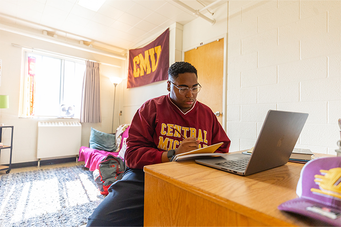 A student sitting at a desk writing on a paper in an Emmons Hall dorm room.