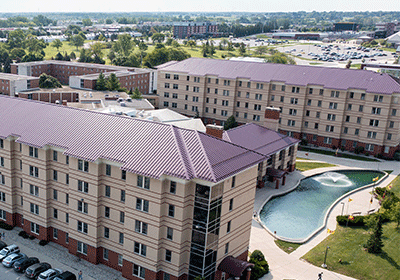 Aerial view of Fabiano Hall on the campus of Central Michigan University, captured from a drone. The residence hall is surrounded by lush green grass and trees, with clear blue skies above and a fountain out side.